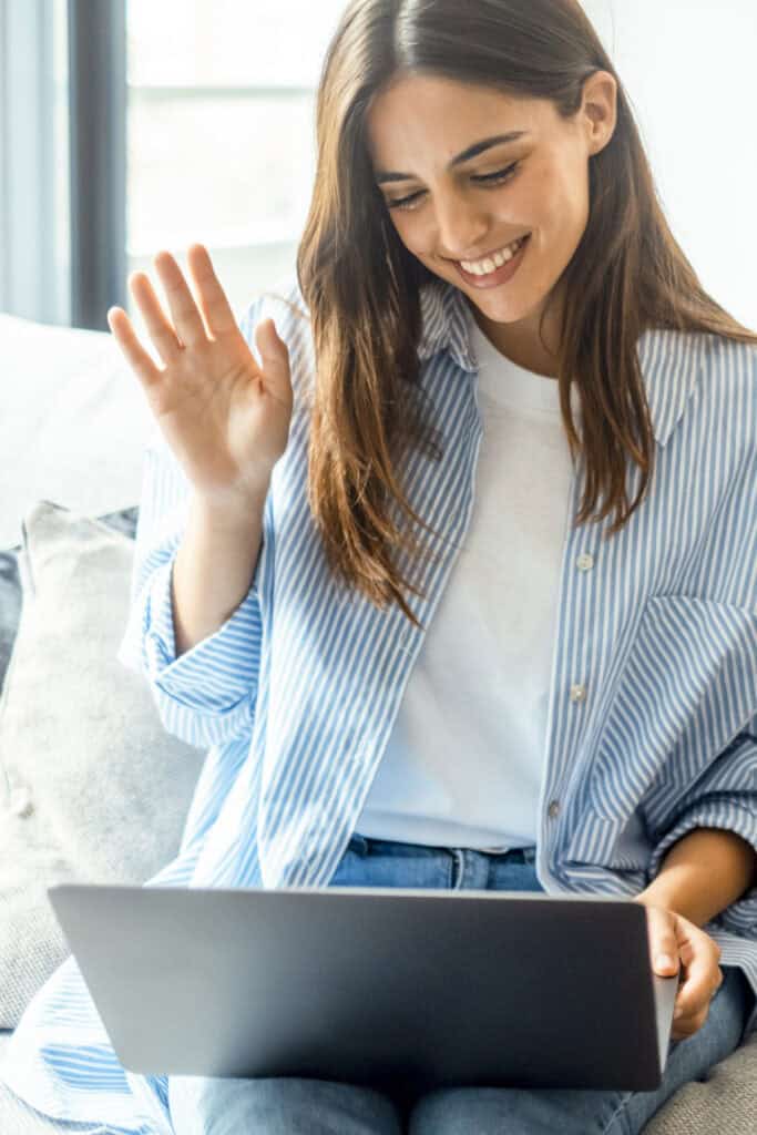 Woman smiling and waving at her laptop during a video call, illustrating online anxiety therapy.